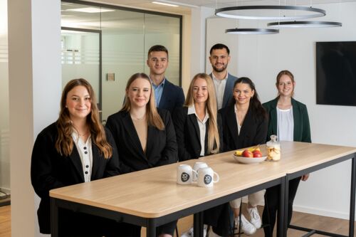 Gruppenfoto der neuen Auszubildenden der Lampe & Schwartze Group: Aylin, Marie-Louise, Shanice und Melissa sitzen auf Barhockern an einem hohen Tisch, Sarah steht daneben und Erik und Ghenadie stehen mittig hinter den Mädchen.